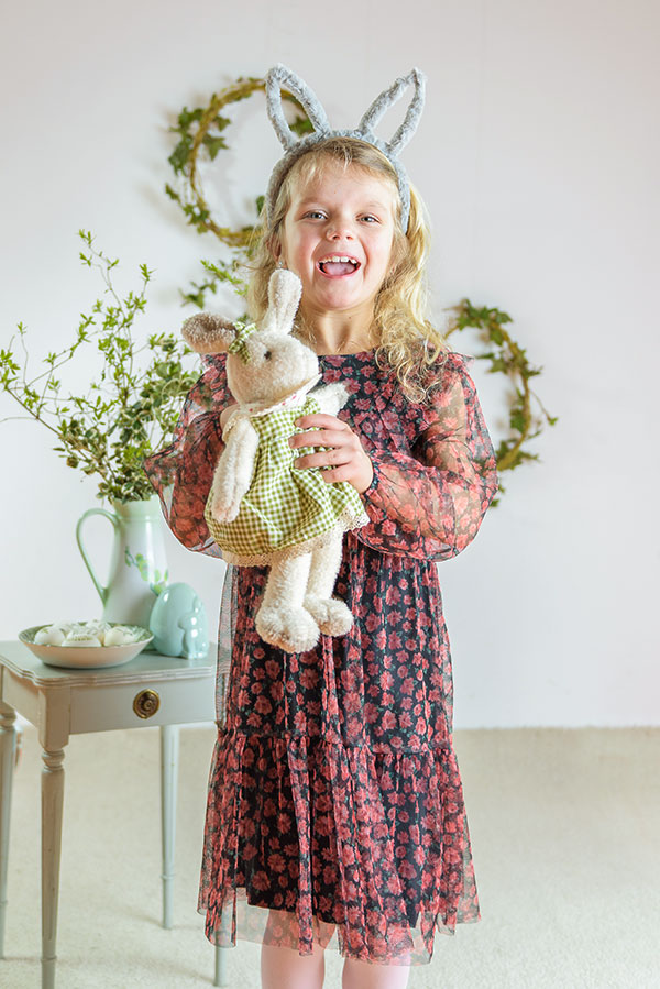 Wesoła dziewczynka w uszach króliczka, trzymająca pluszowego zajączka, z radością witająca wiosnę w kwiecistej sukience, na tle delikatnej wiosennej dekoracji.