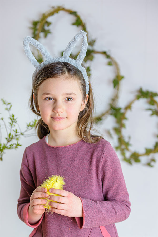 Urocza dziewczynka w wiosennym nastroju, z uszami króliczka i trzymającą żółte pisklę, z wiankiem zielonych listków tworzącym subtelne tło.