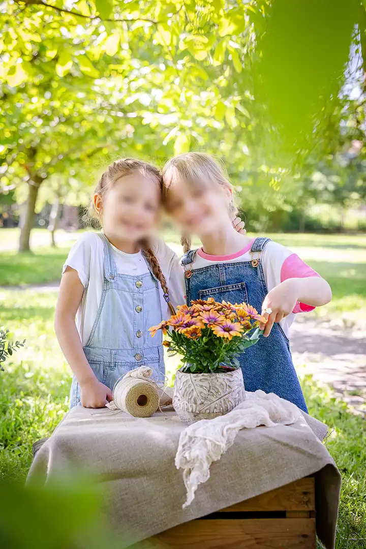 Dwie dziewczynki sadzą kwiaty w doniczce pośród zieleni. Na stoliczku przed nimi są są różne akcesoria ogrodnicze.