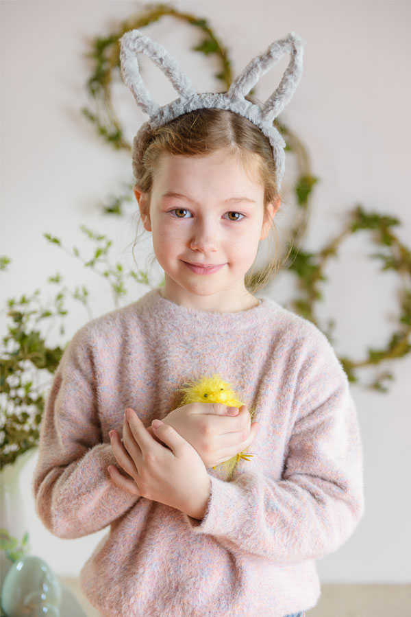 Dziewczynka w uszach króliczka delikatnie trzyma małego, żółtego pisklęcia, otoczona wiosenną roślinnością, co przywołuje nastrój radosnego i beztroskiego Święta Wielkanocnego.