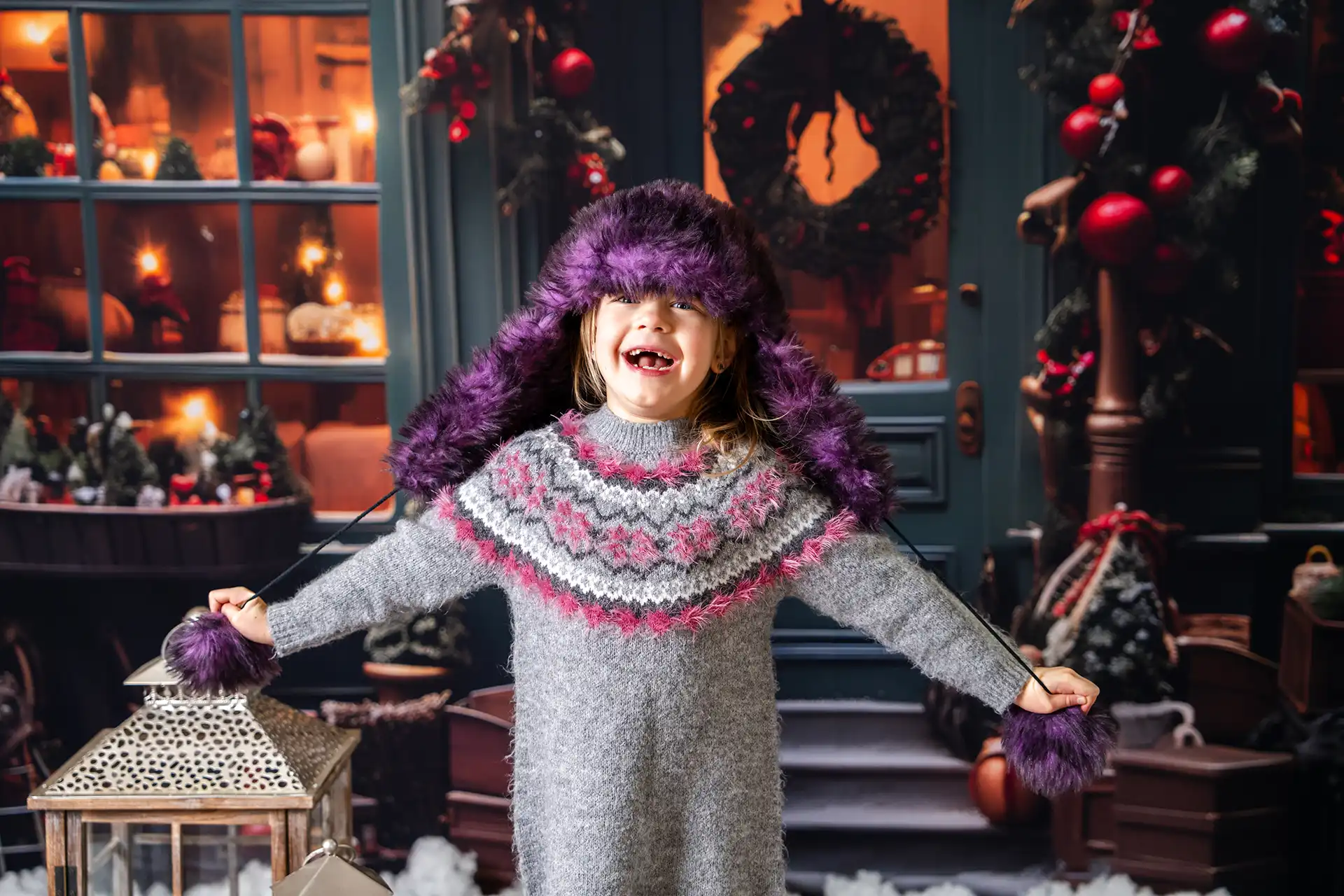 Uśmiechnięta dziewczynka w ciepłym, szarym swetrze i kolorowej zimowej czapce, rozpościera ręce w radosnym geście, stojąc przed świątecznym wnętrzem z ozdobionymi choinkami.